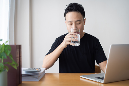 Asian young man in black shirt drinking a water in drinking glass while taking a break. Work life balance and health care in working people in modern lifestyle. Thirsty businessman drink a water.