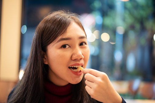 Asian woman enjoy eating French fry or potato chip in the fast food restaurant at night. Unhealthy eating behavior in young adult. Often eating the junk food is not good for health.