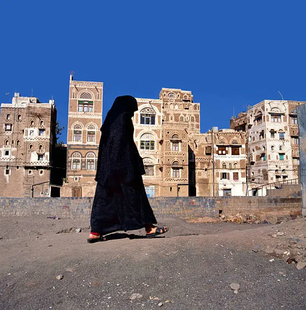 A veiled Muslim woman walks on a Sana a street, Yemen.At background typical Yemen houses.