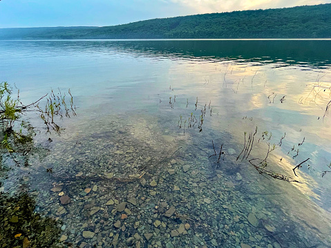Rocks on the shoreline of a tranquil lake on a gloomy overcast day surrounded by forested hills and mountains reflected in the water