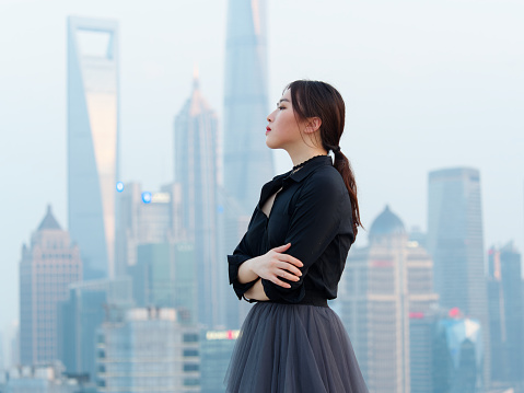Beautiful young Chinese woman in black arms crossed on top of mansion roof with blur Shanghai Bund landmark buildings background with dusk light. Emotions, people, beauty and lifestyle concept.