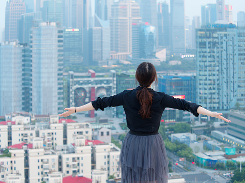 Rear view of beautiful young Chinese woman in black opening her arms on top of mansion roof with blur Shanghai Bund landmark buildings background. Emotions, people, beauty and lifestyle concept.