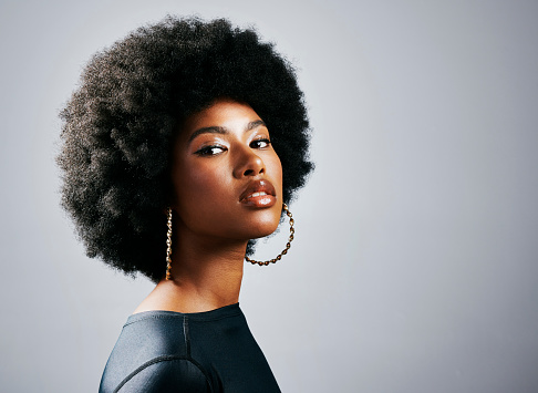 Closeup of a young, stylish, and edgy black woman standing or posing against a grey studio background alone. One confident, trendy and sassy female with an afro looking powerful and wearing makeup