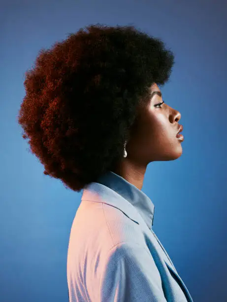 Photo of Profile of fierce, afro and retro model with attitude, perfect skin and a cool, funky and trendy hairstyle. A powerful portrait of intense, proud and fashionable woman against blue studio background