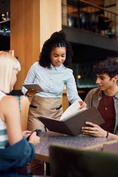 istock Happy black waitress showing a menu to guests in restaurant. 1411158485