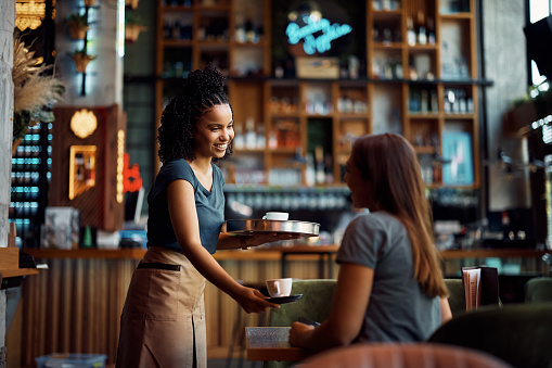 Happy African American waitress serving coffee to female customer while working in a cafe.