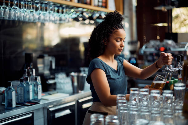 Young black waitress pouring beer draft beer while working at bar counter. African American female barista pouring beer from a beer tap while working in pub. bartender stock pictures, royalty-free photos & images