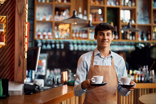 Happy waiter serving coffee while working in a cafe and looking at camera.