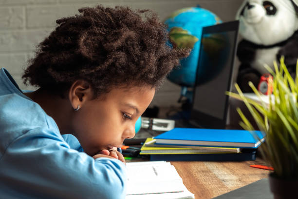 Sad bored African-American girl doing homework at home at her desk,reading a textbook.Back to school concept.School distance education,home schooling,diverse people stock photo