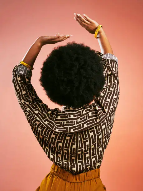 Young woman with hands raised high, happy and dancing listening to music and having fun. Portrait from behind of a funky, attractive and stylish female with afro hair isolated on a brown background