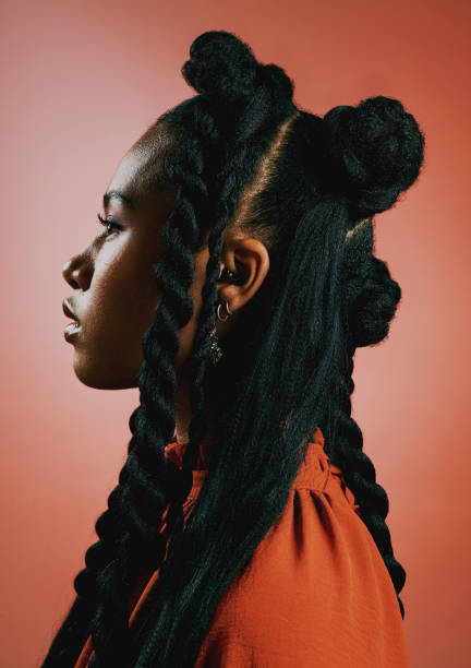 joven negra vanguardista con peinado trenzado de moda sobre un fondo de estudio naranja. perfil de una mujer afroamericana segura, seria y elegante con personalidad audaz y actitud fresca - trenzas fotografías e imágenes de stock