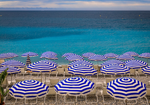Pebble beach along Promenade des Anglais with beach umbrellas and chairs with the turquoise water of the Mediterranean Sea in Nice, Cote d'Azur France