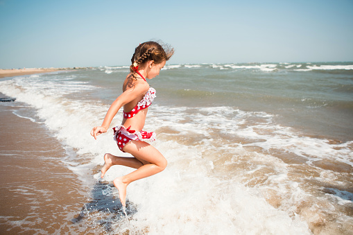 A little girl in a red swimsuit is playing on the beach with a sea wave - jumping, running, having fun. Swimming, traveling, playing with water