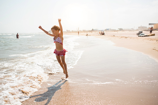 A little girl in a red swimsuit is playing on the beach with a sea wave - jumping, running, having fun. Swimming, traveling, playing with water