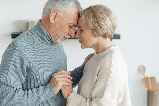 Profile view of elderly married couple hugging, standing together at home. Gray haired man care and support his wonderful woman, while she standing nearby with closed eyes
