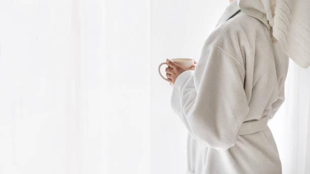 Woman in bathrobe standing near window with coffee cup