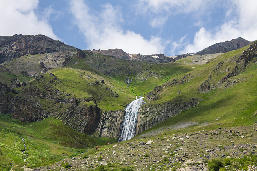 The high-altitude waterfall Terskol flowing from the top of a cliff among alpine meadows in a valley in the Elbrus region in the North Caucasus on a cloudy summer day