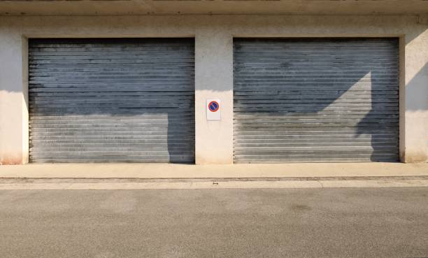istock Large roller shutter garage doors on a concrete facade with a no stopping sign in the middle. Cement sidewalk and asphalt road in front. 1411177223