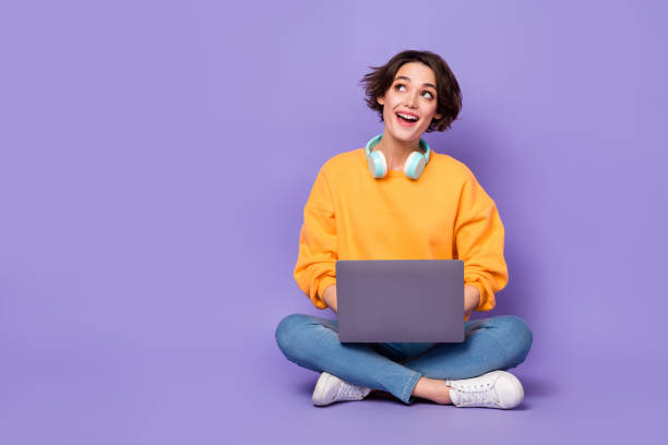 魅力的なガーリー陽気な好奇心旺盛な女の子の肖像画は、バイオレットライラック色の背景の上に隔離されたコピースペースを考えて蓮のポーズをとって座っている - women people internet laptop ストックフォトと画像