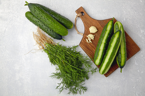Peeled fresh cucumbers on a brown wooden cutting board with a bunch of dill and garlic on a light gray concrete table. Cook at home. Top view with copy space