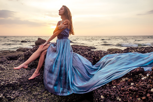 gorgeous style brunette young woman in blue chameleon dress long train standing on a rock near the sea, sand tropical on a rock.beautiful mermaid lonely melancholy waiting freedom and freelancing.