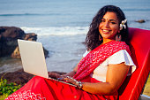 remote working concept dream job. indian business woman in red stylish sari ,curly hairstyle,toothy white smile and flower in hair working with a laptop while sitting in a summer cafe by sea
