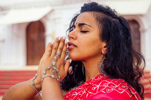 Young Indian woman in traditional sari red dress praying in a hindu temple goa india Hinduism.girl performing namaste gesture catholicism Delhi Street holi festival.om yoga meditation female model.