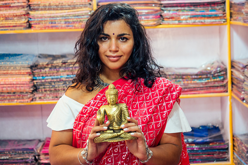 business lady indian seller tradition red sari souvenir shop buddha shiva figurine yoga meditation.girl in india in the religion store.beautiful woman with bijouterie jewelry earrings Delhi Bazaar.
