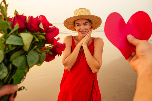 hands of a man giving a heart shaped cardboard valentine and a bouquet of roses to his beloved woman surprised and beautiful woman, in red dress and straw hat on the beach by the sea The 14th February..