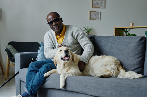 Portrait of African blind man in dark glasses resting on sofa in living room together with his guide dog