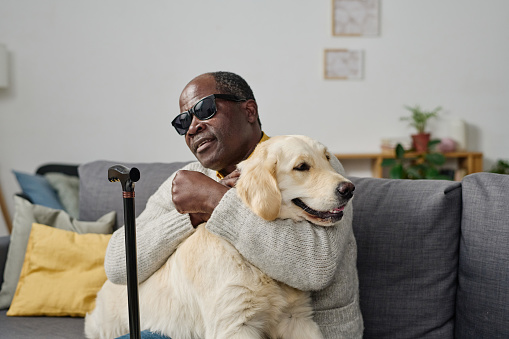 African senior man in glasses with bad sight embracing his individual guide dog while sitting on sofa at home
