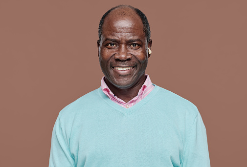 Portrait of African senior man with disability with hearing aid smiling at camera against beige background
