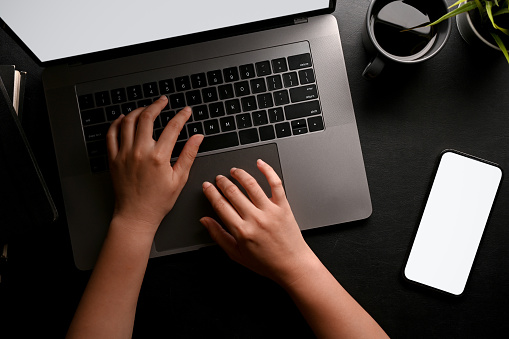 A female hands or businesswoman typing on notebook laptop keyboard, smartphone white screen mockup and decors on black workspace background. top view