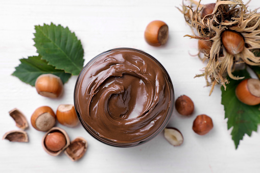Jar with tasty chocolate spread, hazelnuts and green leaves on white wooden table, flat lay
