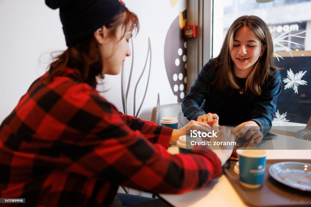 Teenage girls enjoying street food and coffee inside Two teenage girls sitting by the window in cafe and enjoying pastry with coffee in paper cups, street food and drinks Cafe Stock Photo