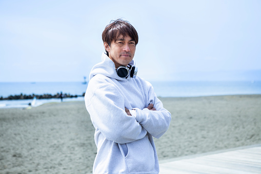 A 40s Japanese man standing on a beachside terrace.
He wears a white hoodie and headphones.