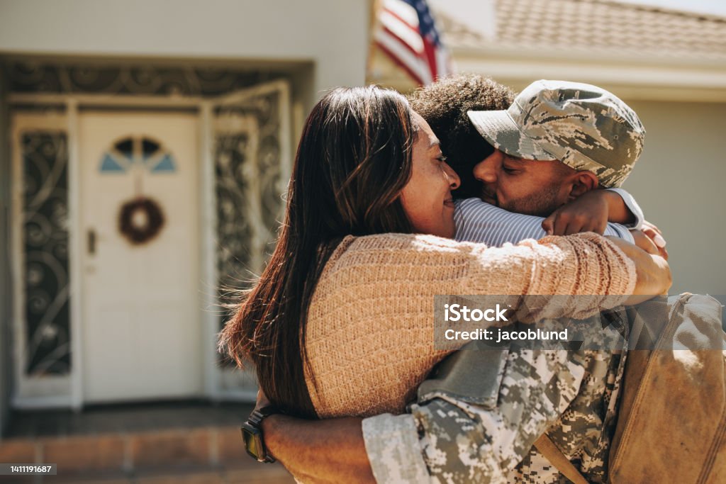 Military serviceman reuniting with his family at home Military serviceman reuniting with his family after deployment. Soldier embracing his wife and daughter after returning from the army. Military man receiving a warm welcome from his family at home. US Veteran's Day Stock Photo