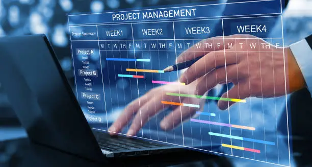 Photo of Project manager working on laptop and updating tasks and milestones progress planning with Gantt chart scheduling interface for company on virtual screen. Business Project Management System.