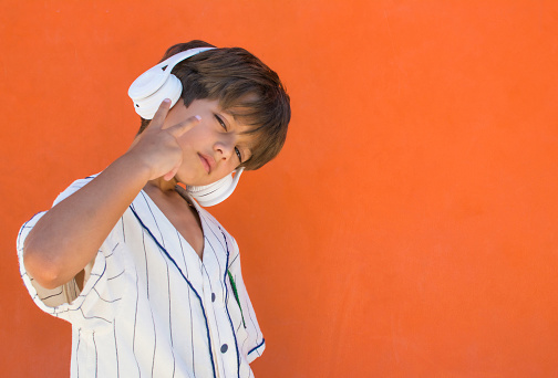 Little boy of 7s listening to music with white headphones.
Cheerful child with funny gesture listening to music with white headphones on orange background