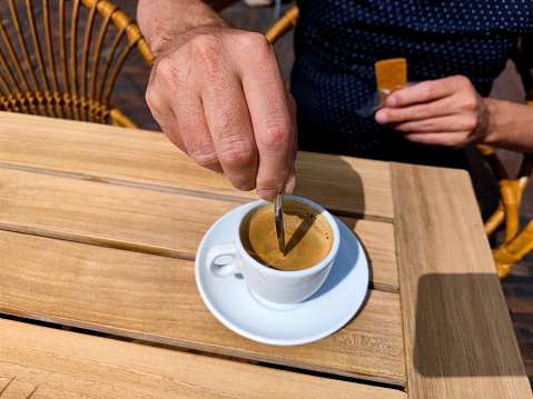White caucasian male hand stirs a freshly brewed cup of coffee on a patio terrace on a wooden table. There are no recognizable people or trademarks in the shot.