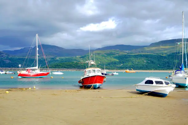 Boats beached at low tide on the beach with the mountains of Cader Idris and the railway bridge of Barmouth in Wales , July 2022.
