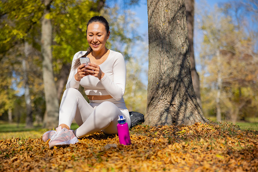 Cheerful Asian woman in casual sports clothes sitting on dry leaves by the tree in the park, texting on smart phone and relaxing outdoors