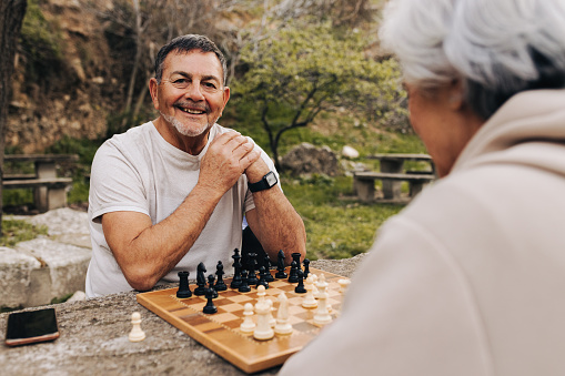 Cheerful senior man playing chess with his wife in a park. Happy elderly couple spending quality time together after retirement. Mature couple enjoying themselves outdoors.