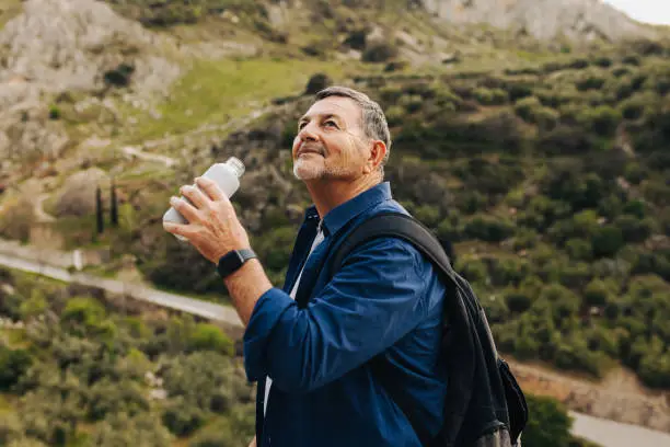 Mature man drinking some water while standing on a mountaintop. Adventurous senior man enjoying a leisurely hike outdoors. Elderly man enjoying recreational activities after retirement.
