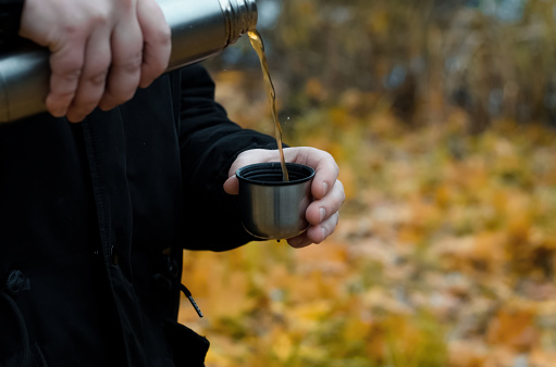 Man pours a hot drink from a thermos into a mug outdoors. Close-up male hands holding a hiking flask against the background of an autumn forest
