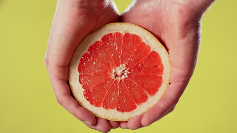 Hands holding grapefruit and squeezing the juice out isolated on yellow studio background. Tropical citrus fruit known for its sour to semi sweet taste. Refreshment high in vitamins and fiber