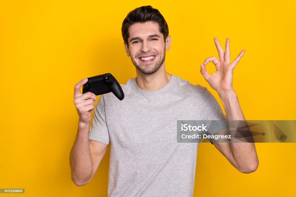 Photo of funny brunet millennial guy playstation show okey wear grey t-shirt isolated on yellow color background Photo of funny brunet millennial guy playstation show okey wear grey t-shirt isolated on yellow color background. Joystick Stock Photo