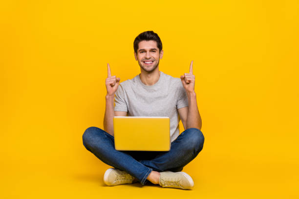 Full length photo of funny brunet millennial guy sit with laptop index up wear t-shirt jeans shoes isolated on yellow background stock photo