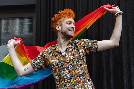 Young man holding a rainbow flag in front of the black wall. Alternative looking man dancing and waving with LGBT flag as symbol of love
