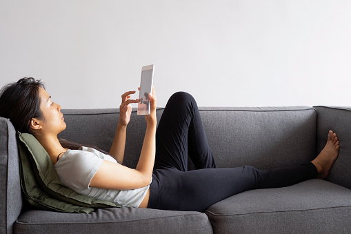 Full body side view of a young Asian woman doing business on a digital tablet while laying on a sofa